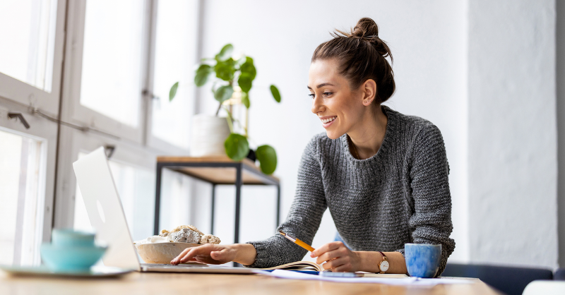 Photo of a woman sat a desk with a pen in her hand. She is looking at a laptop.