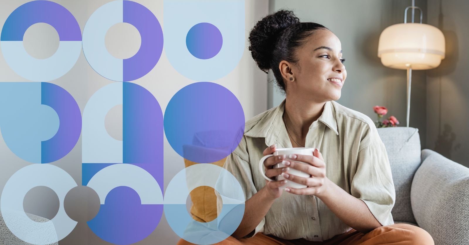 A smiling and confident care worker sits cross-legged on a sofa looking out the window, holding a mug of tea.